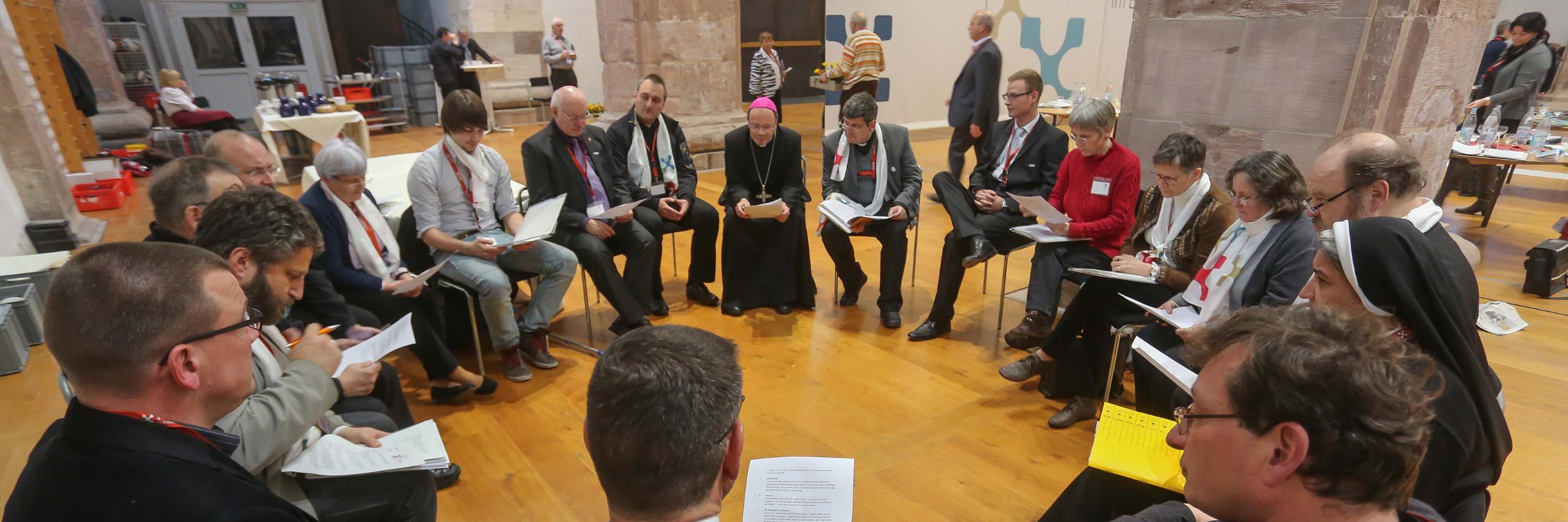 Viele Frauen und Männer sitzen gemeinsam mit Bischof Ackermann im Kreis. An der Wand im Hintergrund sind die Zeichen für die Synode zu sehen.