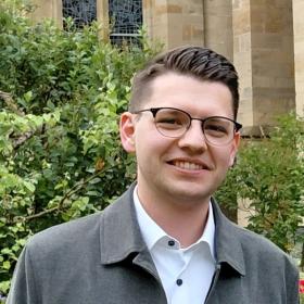 Die künftigen Diakone: Manuel Hauth und die ständigen Diakone Torsten Wahl und Raphael Collinet (v. l.n.r.) (c) AxelBerger/Bistum Trier Drei Männer stehen lächelnd im Grünen; Im Hintergrund die Liebfrauenkirche
