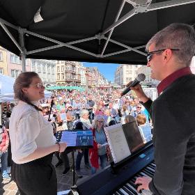 Eine Frau und ein Mann haben Mikrofone in der Hand, der Mann spielt Keyboard und sie stehen auf einer Bühne vor der sehr viele Leute auf einem Platz stehen.